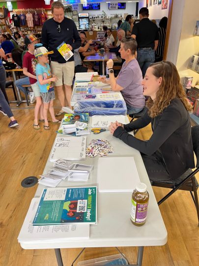 The author of the article sits in the foreground at a folding table covered in stickers and flyers promoting the freedom to read. She is a young woman with curly light brown hair, turned in profile to the camera and smiling. In the background, a family with an adult and two children is walking up to the tables.
